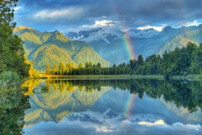 A calm lake reflects surrounding mountains, trees, clouds, and a rainbow in the sky.