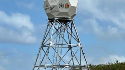 A geodesic dome-shaped weather radar tower stands on a metal framework against a background of blue sky and clouds. Logos are visible on the dome's surface.