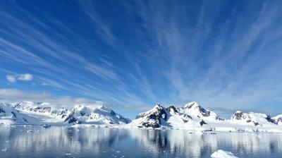 Snow-covered mountains and glaciers are reflected in calm Antarctic waters under a blue sky with wispy clouds.