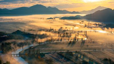 Aerial view of a misty landscape featuring meandering rivers, fields, and distant mountains at sunrise.