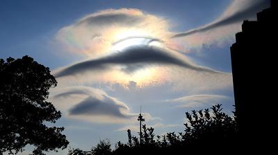 Clouds surrounding the sun with iridescent colors, silhouetted by trees and a building, set against a blue sky.