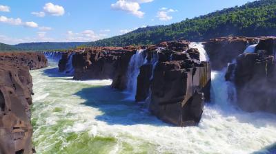 Waterfalls cascade over rocky cliffs into a turbulent river under a blue sky with scattered clouds, surrounded by a forested landscape.