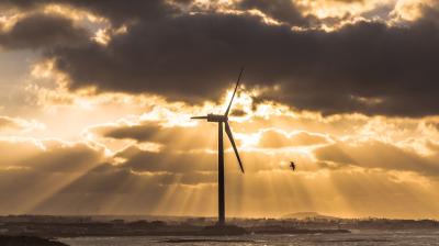 A wind turbine stands by the water at sunset, with clouds and sun rays in the sky and a bird flying nearby.