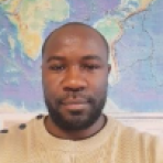 A person with a beard is in front of a world map, wearing a light-colored sweater.