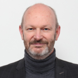A person with a beard, wearing a dark blazer and gray turtleneck, against a white background.