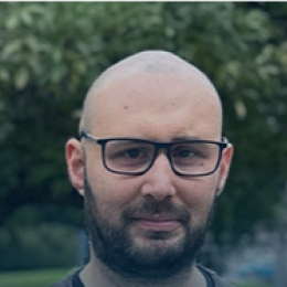 A person with glasses and a beard is outdoors with trees in the background.