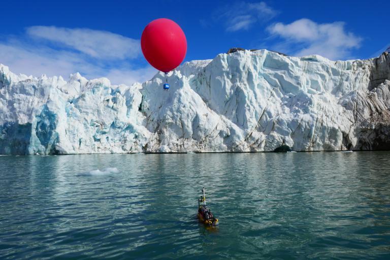 A glacier with a floating red balloon above it and a small, partially submerged device in the water below, set against a blue sky with scattered clouds.