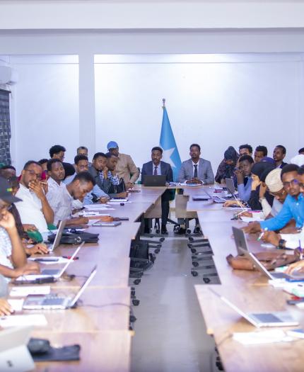 A large group of people in a meeting room, sitting at a long table facing forward. A flag is positioned at the end of the table. Many have laptops open.